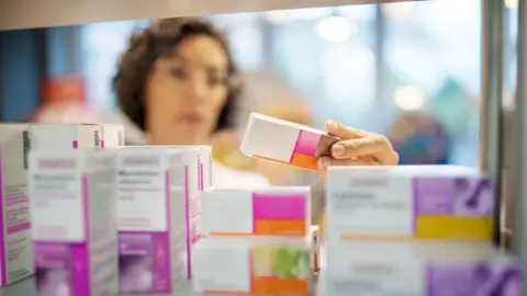 A woman, not in focus, handles different packets of tablets in a pharmacy setting