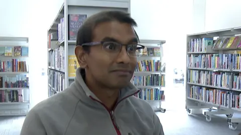 Doctor Kamlesh Sreekissoon is sat in a library with shelves filled with books behind him. He is wearing a fleece and glasses. 