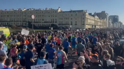 Allison Ferns/BBC A shot from above of dozens of people dressed in brightly coloured outfits running a marathon on Brighton seafront, with cream coloured buildings behind them