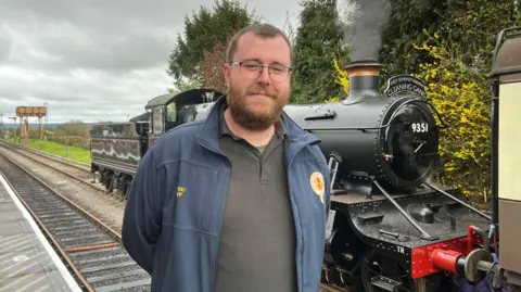 A bearded man wearing glasses and a blue jacket is stood in front of a black locomotive on a railway line.