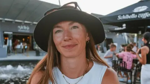 Megan Chapple is a woman in her mid 20s. She is smiling and has green eyes. She is wearing a black sun hat, with sun glasses places on top and a white top with gold necklaces and earrings. She is sat in a beer garden. 