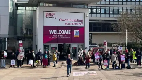 A group of people are standing outside Sheffield Hallam University with pink and white placards. In the background is a large entrance with a pink and white sign