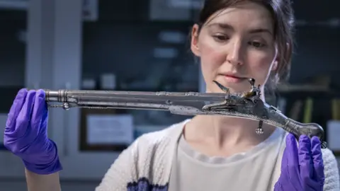 National Museums Scotland / PA Curator Ella Paul holds one of the Jacobite pistols aloft. She is wearing a white top and white cardigan and purple protective rubber gloves.