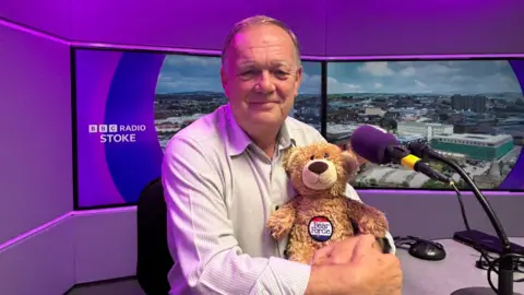 A man sitting in a radio studio with tv screens behind him is looking at the camera and holding a bear. The man is wearing a light-coloured shirt and has a purple microphone in front of him.