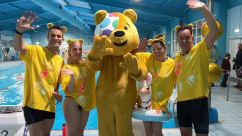 BBC Pudsey Bear centre with the Radio Solent and BBC South swimmers either side and a swimming pool in the background.