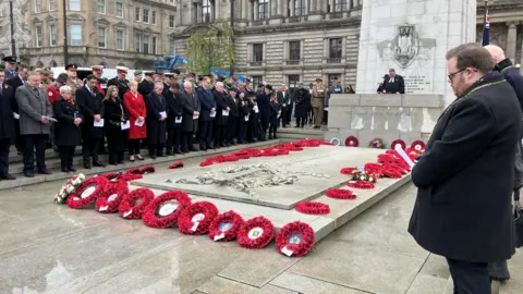 Wreaths around the cenotaph in George Square. Mourners dressed in black are standing around the side of the image.