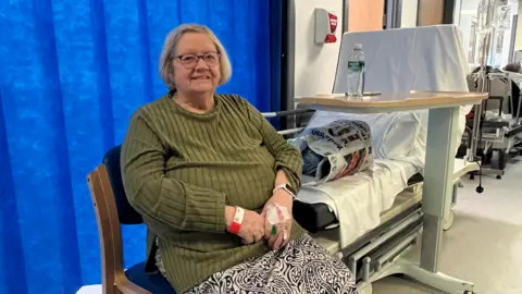 An 80-year-old woman in a green jumper and black and white patterned skirt is sitting on a chair in a hospital corridor. She is next to a bed that has a carrier bag on it with her possessions. She has short grey hair and is wearing glasses. She has a blue curtain behind her.
