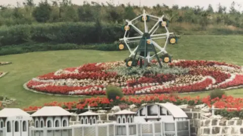 Helen Sherwin A large floral display in red and white on a lawned hillside, with a small ferris wheel in the centre.