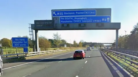 A blue sign on a board over a motorway indicating a slip road for the M55 towards Blackpool and Preston