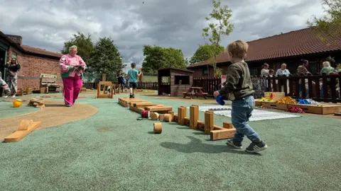 A woman wearing pink trousers and a pink shirt and cardigan is walking across an outdoor playing area at the centre, where a young boy wearing trainers, blue jeans and a green jumper is seen walking towards an assortment of wooden play items.
