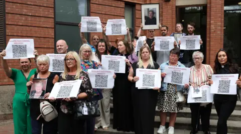 PA Media A group of women and men stand on steps outside a red-brick courthouse. Each of them holds a sheet of paper with a QR code and the words "Regulate our funeral industry" written on them. Some of them are also holdings photographs of loved ones