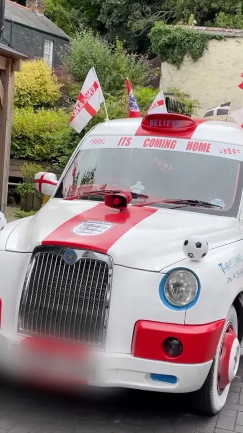 A white and red car which has "its coming home" written in red letters. There are english flags and footballs over the car. The number plate is blurred.