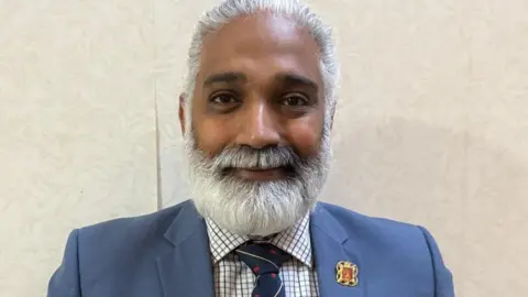 Lancashire County Council Dr Sakthi Karunanithi wearing a blue suit with a checked shirt with a large white beard. He stands in front of a wall with cream wallpaper. He is smiling.