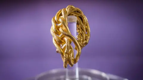 A close-up of the fragment of gold arm-ring. It has eight strands of gold rods which have been plaited together. It hangs on a clear stand in front of a purple background.