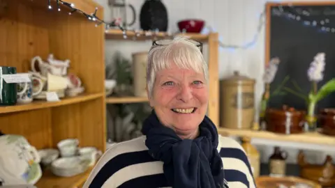 Lyn Osbaldeston, pictured in her shop in Cockermouth. She has short white hair and blue eyes. She is wearing a navy blue and white top, and a navy blue pashmina knotted at the neck.