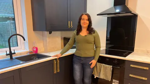 HMO landlord Alice Garnett in green top and blue jeans smiles in newly finished kitchen of her third HMO property in Derby. Kitchen has black cupboards with gold handles and a matte finish, with wooden floor.