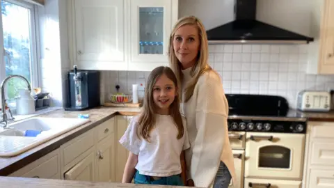 An eight year old girl with brown hair wearing a white t-shirt stands next to her Mum who has blonde hair and is wearing a cream jumper in their kitchen.