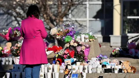 A woman in a bright pink coat and jeans seen facing rows of flowers, toys and coffee cups from a local business, a memorial for those who were killed and injured during a mass shooting in Tumbler Ridge, British Columbia.