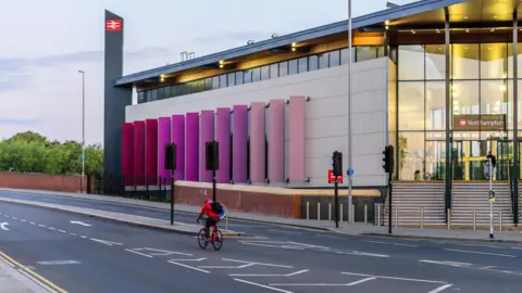 Getty Images A railway station. The building has steps to a glass entrance to the right of picture. The building extends to the left with several long pink and purple decorative boards along the wall. A lone cyclist in red is on the road in front of the station.