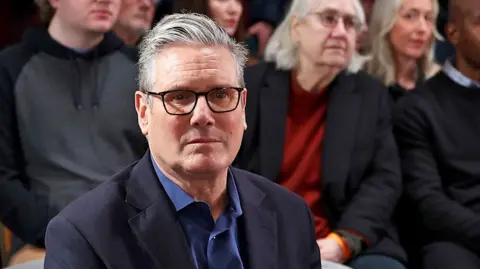 Keir Starmer, wearing black rimmed glasses and an open necked blue shirt, with suit jacket, stares plaintively at the camera. He is sitting in the audience of a press event