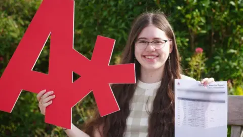 PA Media Gabrielle Morgan has long dark hair and glasses. She is holding up a big red A* sign in cardboard and holding the letter detailing her results. She is smiling and there is a garden behind her. 