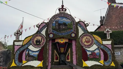 A traditional Derbyshire-style well dressing, featuring an array of colourful designs surrounding a stone well. There is bunting around the well and houses in the background.