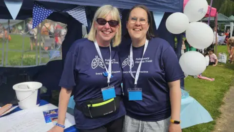 Nicola Lovejoy-Waud Joanne (left) and Nicola Lovejoy-Waud are stood next to a blue stall at an annual fundraising event for Pulmonary Fibrosis, called Picnic in the Park. They are both wearing navy blue T-shirts with the words "Pulmonary Fibrosis Trust, Every breath is precious".