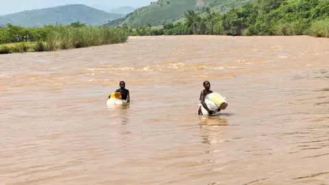 Dua pria yang berjuang melalui perairan cokelat sungai Rusizi yang mencengkeram tas dan kaleng jerry