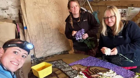 Wendy Greatorex Three women are adding flowers to a huge display board. The display depicts and image of Jesus. They are stood in a workshop.