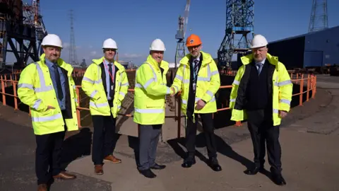 Tim Richardson/UK Docks Marine Services Dan Haslar, senior property manager at PD Ports congratulates UK Docks founder Harry Wilson and his sons Gary, Jonathan and Chris. All five men are wearing yellow hi-vis jackets and hard hats. Mr Haslar's hat is orange. They are standing at the docks.