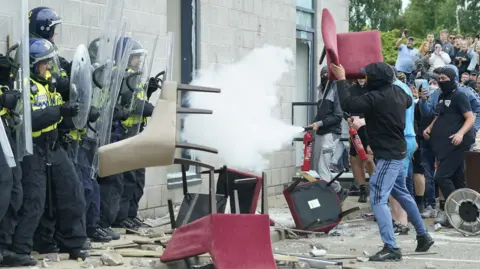 A man wearing blue tracksuit bottoms and a black hooded top throws a red chair at police officers.