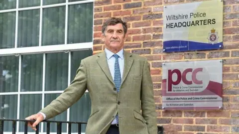 Wiltshire PCC Wiltshire's police and crime commissioner Philip Wilkinson wearing a blue skirt, navy tie and tweed jacket standing outside of Wiltshire Police headquarters, straight-faced and learning against black metal railings. 