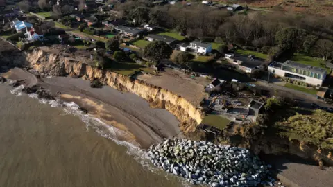 A drone image of a coastline where it is eroding away close to a village. Rock sea defences have been placed at one end of the coastline.