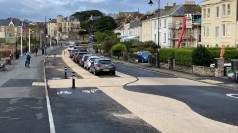 White wiggly lines along the Beach in Clevedon, with cars parked near the sea front. There are homes and cafes along the other side of the road.