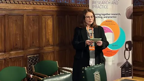 Stevie A young woman with brown hair, wearing a black coat and glasses. She is standing in a room in the Houses of Parliament, in front of a wood-panelled wall and a head-height poster featuring information from the Step by Step charity. She is wearing glasses and  reading from a notepad which she is holding in her hands in front of her. Beside her are two dark green chairs. She has an orange lanyard on which reads "visitor".