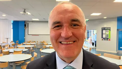 A smiling Michael Allen of Lisneal College is in the school's dining room, with tables and chairs in the background