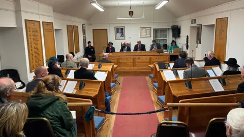 Sark's Chief Pleas in 2024. A group of politicians sitting at wooden benches in front of a desk with six peoiple behind it at the end. The women in the benches are all wearing hats, as is the tradition for Chief Pleas members in Sark. 