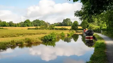 Grum A red, black and cream canal boat on still water with the clouds reflected in it and grassy areas on either side of the water, with a few trees scattered about
