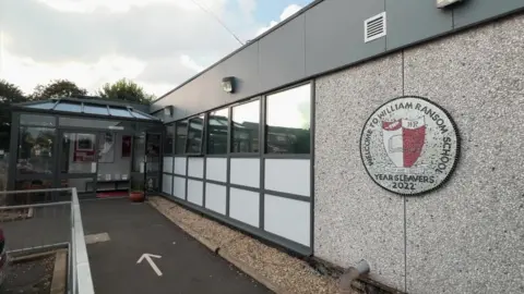 DJ McLaren/BBC The entrance to William Ransom Primary School. A grey building with a school badge by the glass entrance.