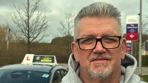 Steve Hubbard/BBC A head and shoulders image of Gerry Harrington. He is wearing a grey hooded top and wearing glasses. He is looking directly into the camera. In the background the top half of a driving instructor car is in shot.