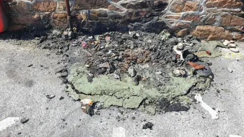 Guernsey Fire and Rescue The remnants of a melted plastic bin on the floor in front of a wall. The remnants include melted plastic, ash and rubble.