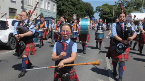 Pacemaker A band wearing red tartan kilts and navy waistcoats. At the front a young girl is leading the band wearing Union flag sunglasses.
