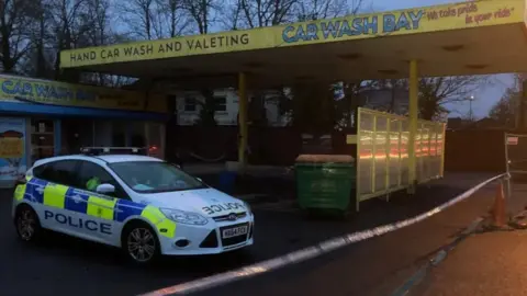 A police car is parked behind police tape in a car wash in fading light