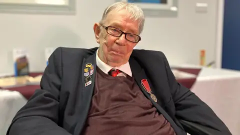 An elderly man sits down and smiles. He has short white hair, a suit jacket with a medal and badges, over a brown top over a white shirt and red tie.