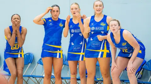 Josh Le Feuvre Five netballers in blue and yellow kit are looking desperately on as their team are defeated in extra time - some have hands up to their mouths in disbelief. They're in an indoor court with pale blue wall and a row of blue fold-up chairs behind them. 