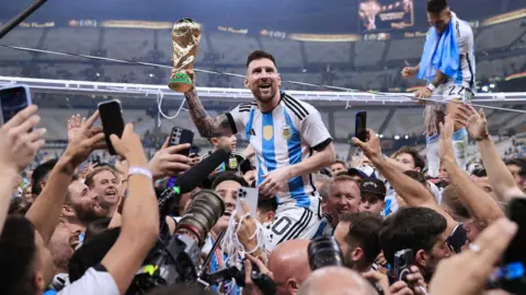 @welloffside/@simonstacpoole A footballer is on the shoulders of another person, grasping a golden trophy. He is surrounded by many other people, some with cameras and phones.