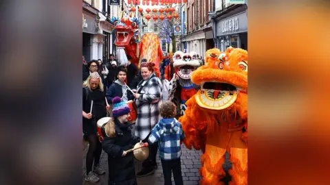Popber and Edward Au A narrow Lancaster street full of shops during a dragon and lion dancing parade to celebrate Chinese New Year. Children and adults are seen playing drums. The people are smiling.