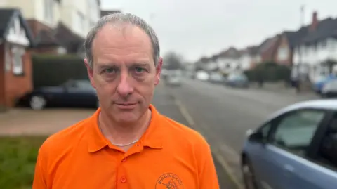 A man in an orange t-shirt which says Birmingham Marlins on it looks at the camera. He stands on the street next to a car.