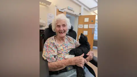 Supplied Joyce pictured at the Barty House Nursing Home in Maidstone with a black stuffed cat. Joyce is wearing a floral patterned t-shirt and is sat in a wheelchair. 
