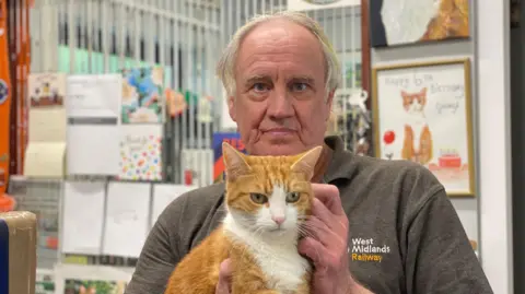 A red and white cat is held by a man in a grey polo shirt with the words 'West Midlands Railway' embroidered on the left side. Behind him are pictures and calendars on the wall, including a drawing of the same cat.